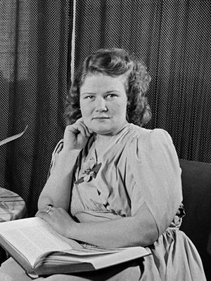 Nainen istuu kirja sylissään, pöydällä leikkokukkia ja radio, 1940-luku. Kuva: <span class="photographer">Väinö Kannisto / Helsingin kaupunginmuseo</span>