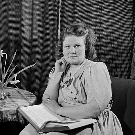 Nainen istuu kirja sylissään, pöydällä leikkokukkia ja radio, 1940-luku. Kuva: Väinö Kannisto / Helsingin kaupunginmuseo