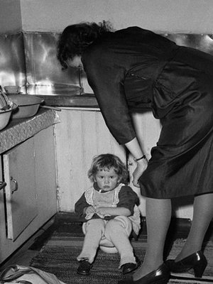 Perhejuhla kotona 1950-luvulla, pieni tyttö istuu keittiössä potalla, äiti seisoo vieressä. Kuva: <span class="photographer">Väinö Kannisto / Helsingin kaupunginmuseo</span>