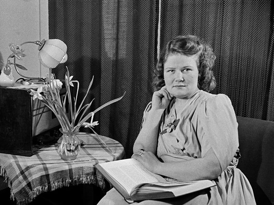 Nainen istuu kirja sylissään, pöydällä leikkokukkia ja radio, 1940-luku. Kuva: <span class="photographer">Väinö Kannisto / Helsingin kaupunginmuseo</span>